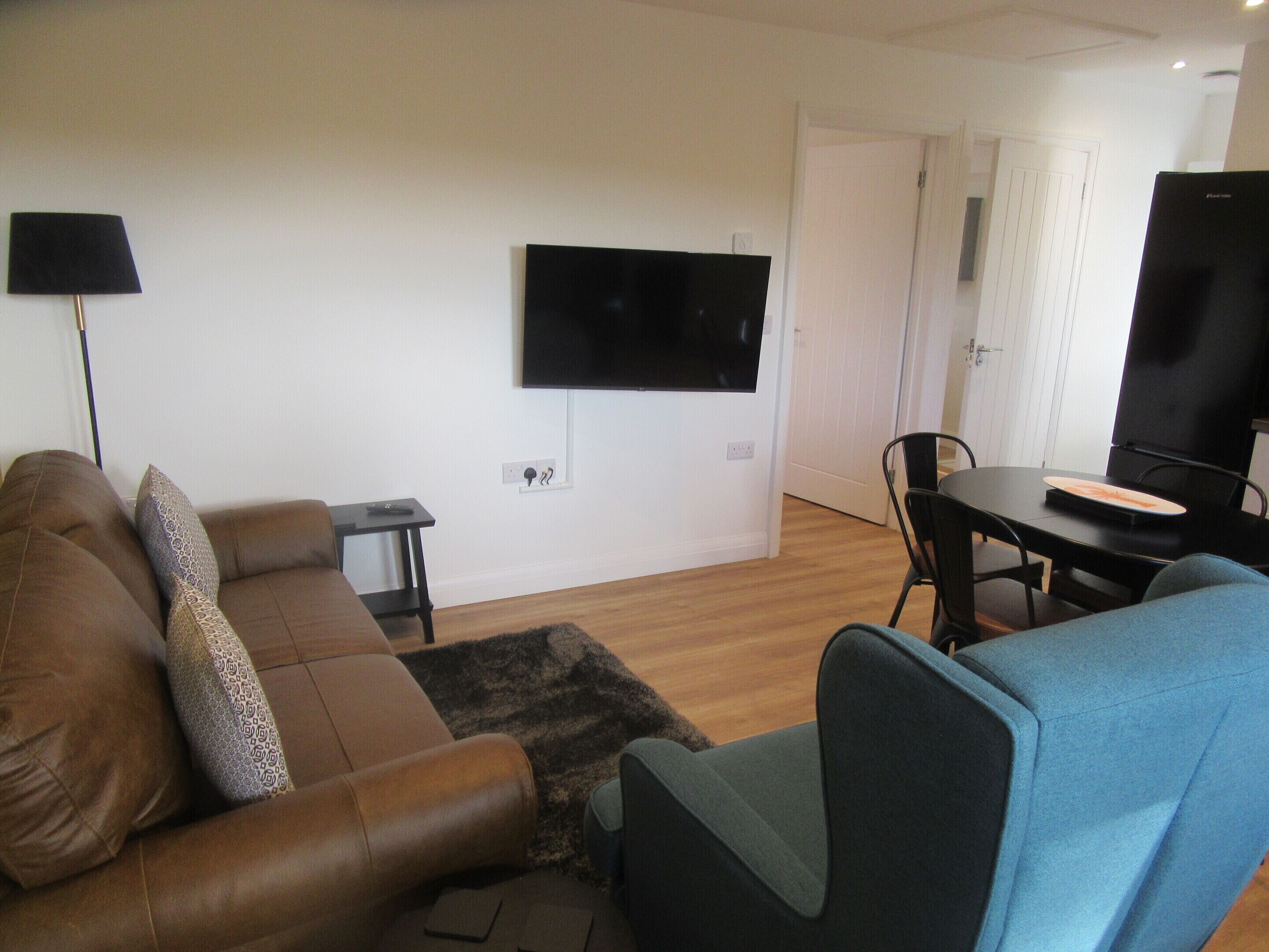 A living room with a brown sofa, a blue armchair, a small table, and a TV mounted on the wall.