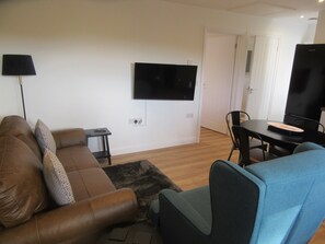 A living room with a brown sofa, a blue armchair, a small table, and a TV mounted on the wall. - Pet Friendly Cottage with Sea Views (Saundersfoot)