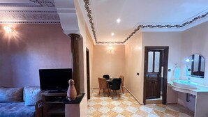 Un salon avec un téléviseur, un vase et un canapé. Il y a également un coin repas avec une table et des chaises. La pièce a un sol à carreaux et une porte qui donne sur une salle de bain équipée d’un lavabo et d’un miroir.
