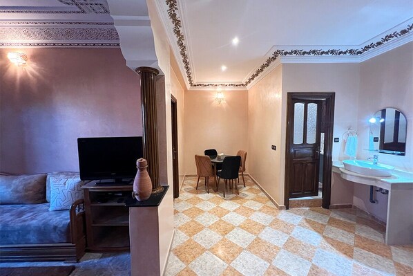 Una sala de estar con televisor, un jarrón y un sofá. Hay una zona de comedor con una mesa y sillas. El piso tiene un patrón de cuadros y una puerta que da a un baño con lavamanos y espejo.