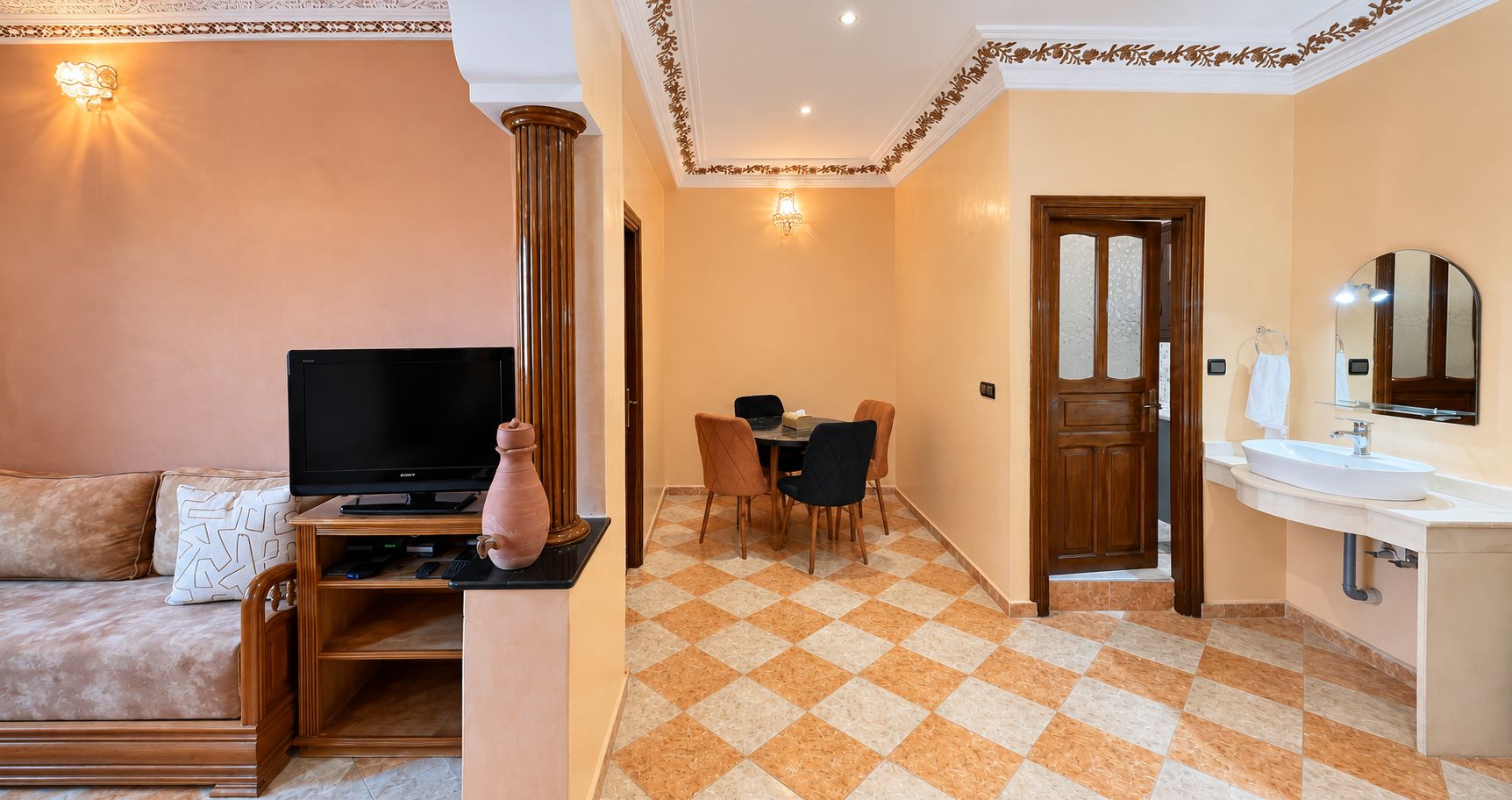Une chambre d’hôtel équipée d’une télévision, d’un coin repas avec des chaises, d’une porte en bois et d’une salle de bain avec un lavabo et un miroir.