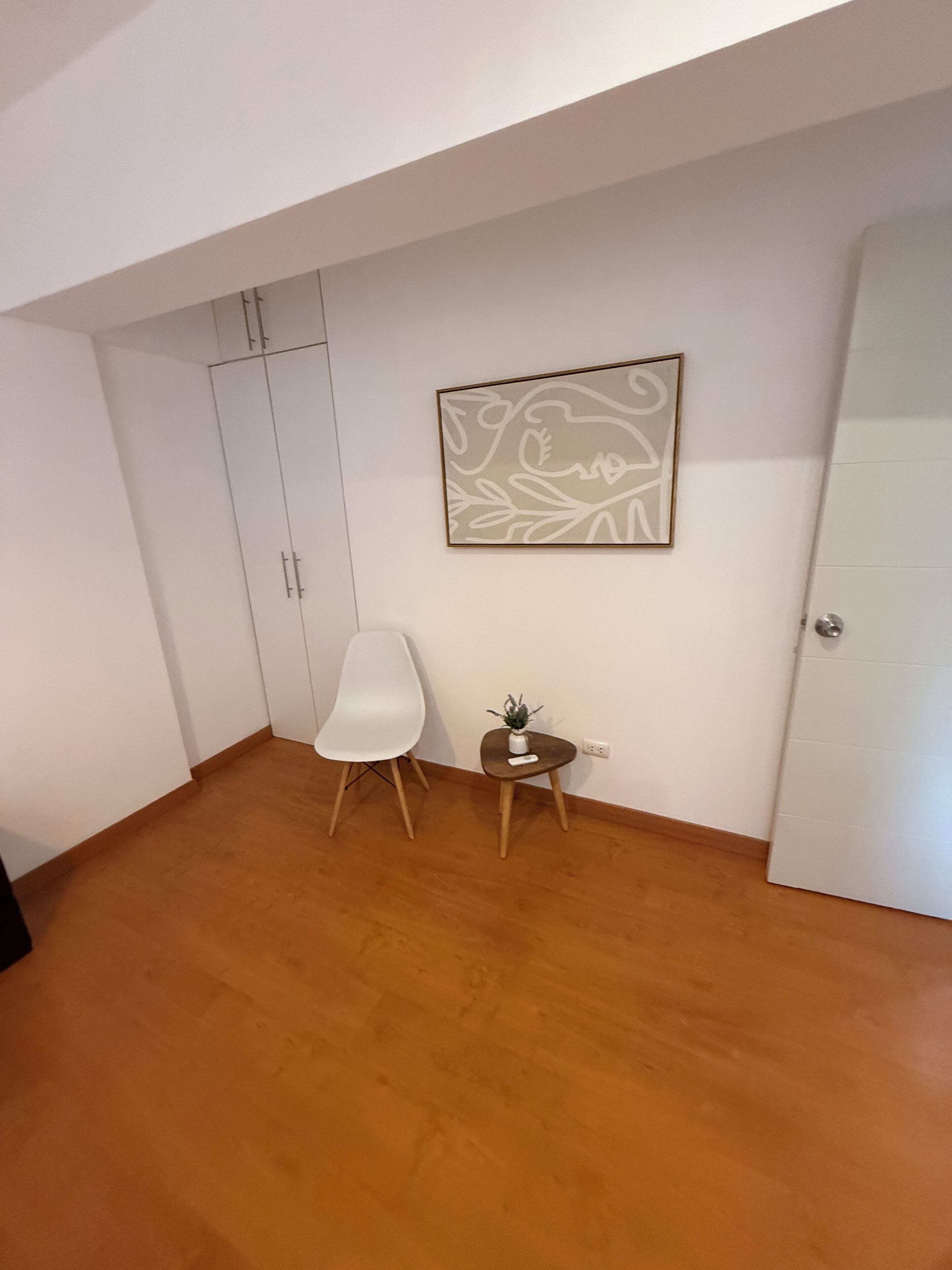 Une pièce avec une armoire blanche, une chaise blanche, une petite table avec une plante et une œuvre d’art encadrée accrochée au mur.