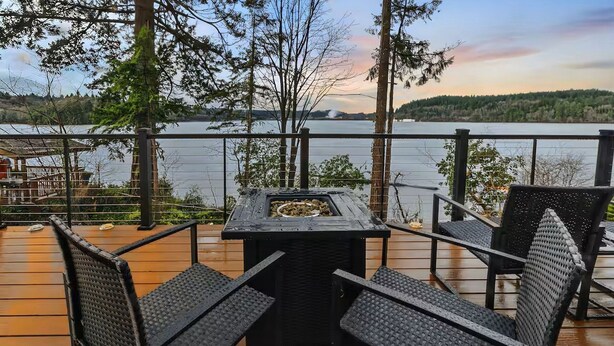 A deck with a table and chairs overlooking a lake, surrounded by trees and a distant shoreline. - Scenic New Built Waterfront Cabin on Hammersley Inlet Olympic Peninsula (Shelton)