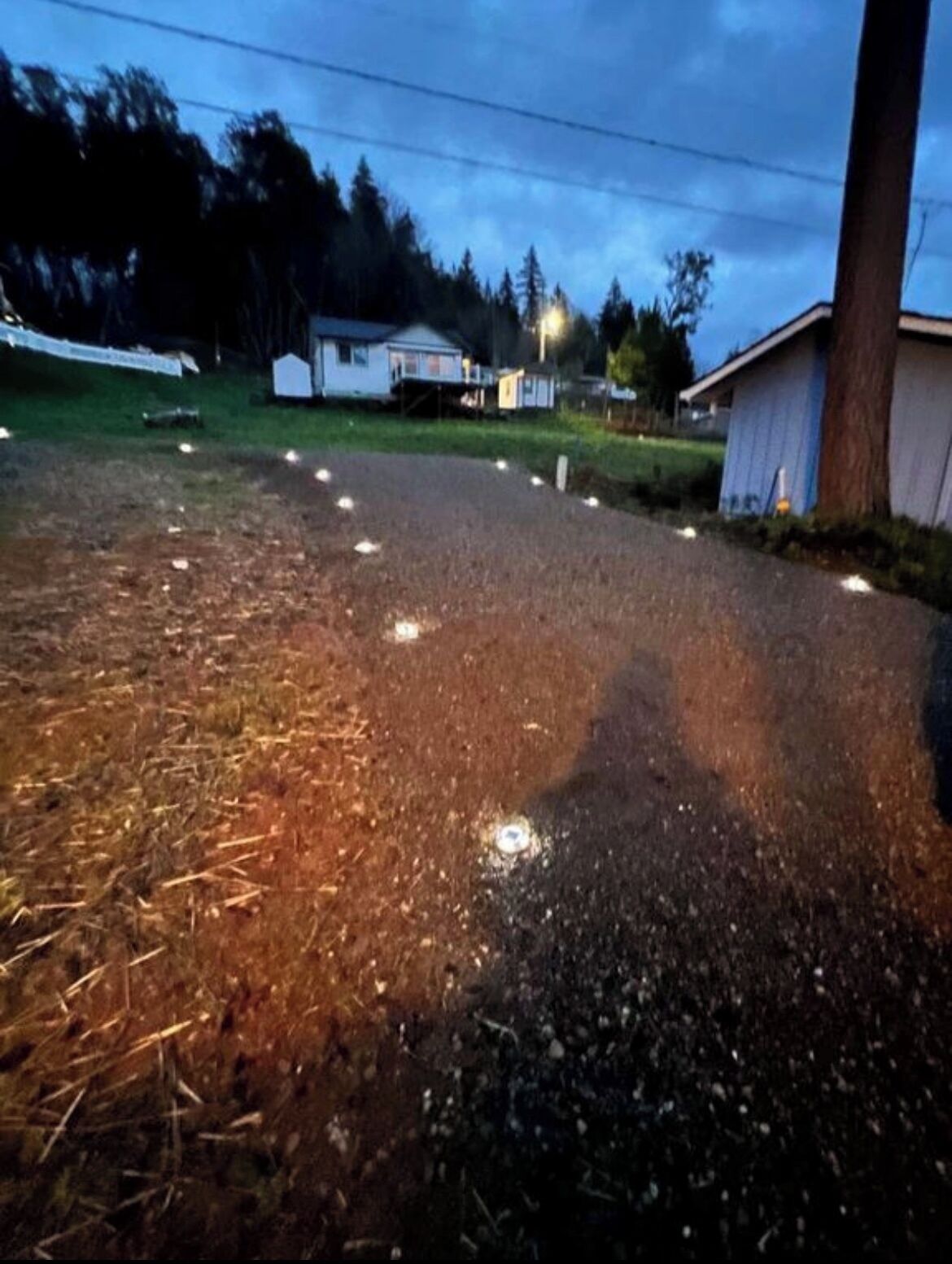 Une allée gravillonnée ponctuée de quelques lampadaires, un petit abri de jardin et une maison en arrière-plan.