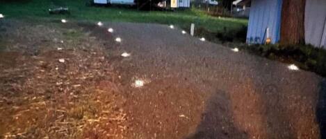 Une allée gravillonnée ponctuée de quelques lampadaires, un petit abri de jardin et une maison en arrière-plan.