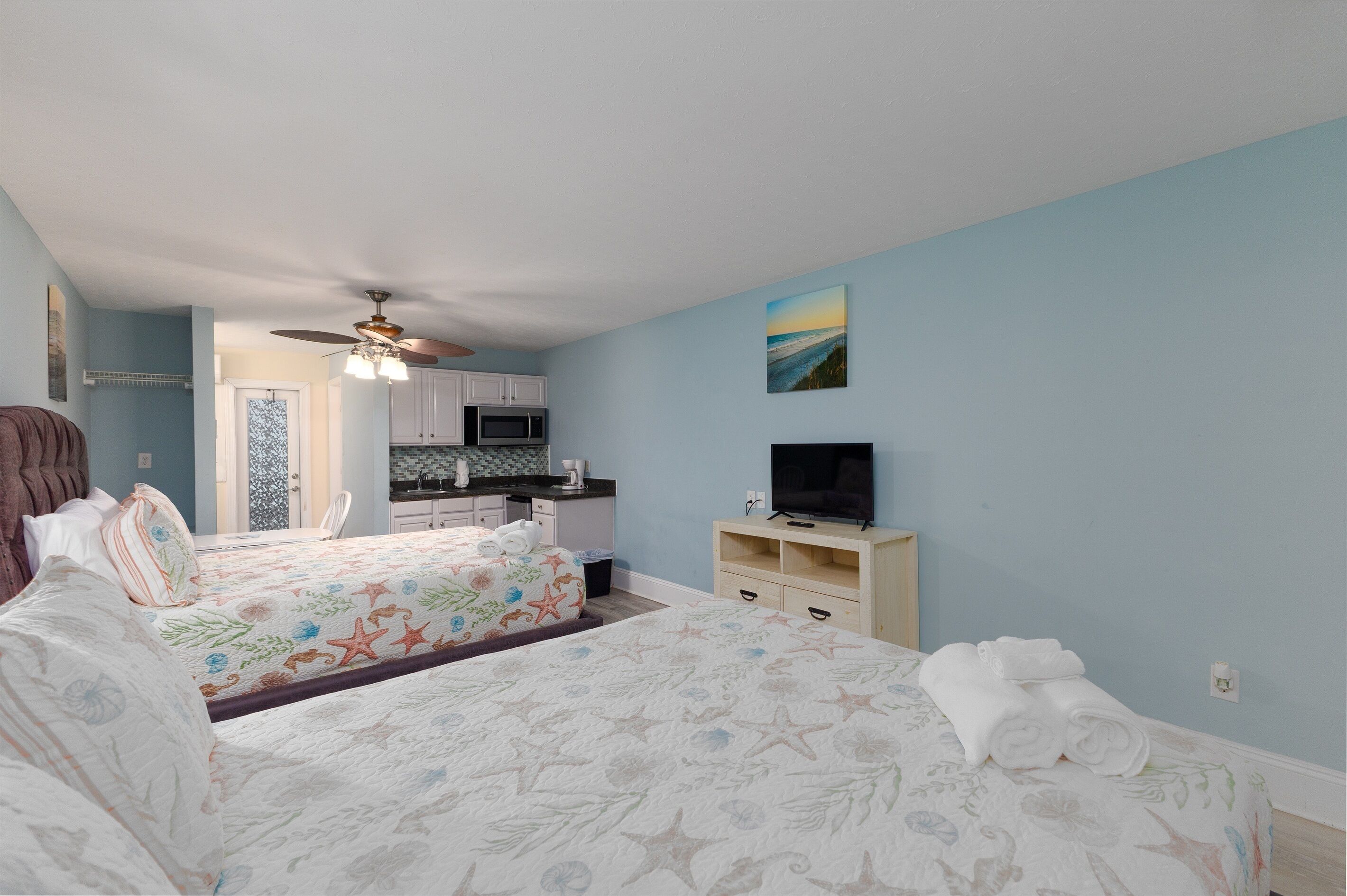 A bedroom with two beds, a TV, and a kitchenette in the background.