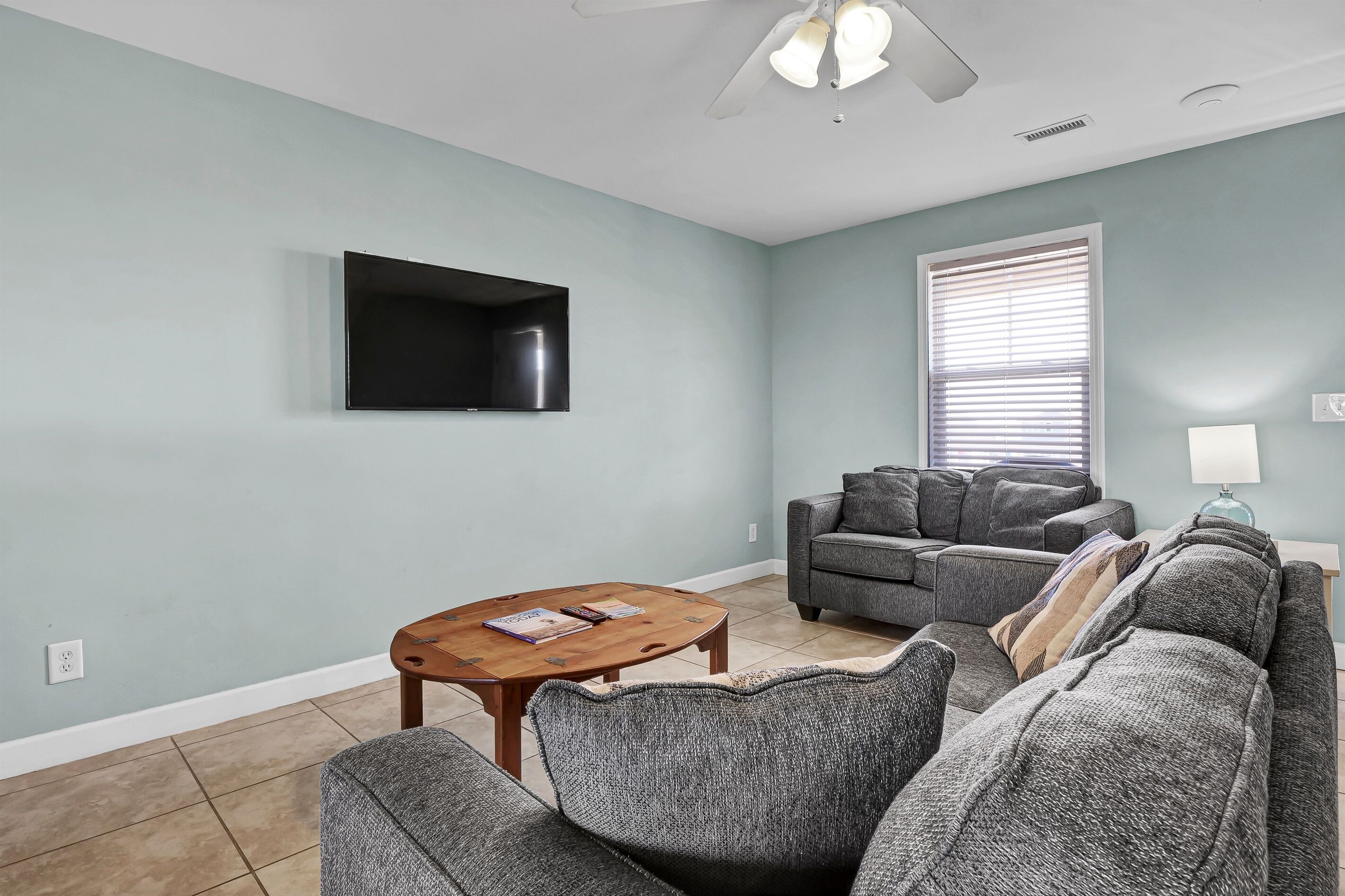 A living room with a flat-screen TV mounted on the wall, a gray sectional sofa, a wooden coffee table with books, and a ceiling fan.