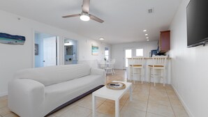 A modern living room with a white sofa, a small white table, and a ceiling fan. There is a kitchen area with white cabinets and a TV mounted on the wall. - Surf City Getaway- Walk to Beach - Seasonal Pool (Surf City)