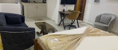 A compact hotel room with a bed, a sofa, a small desk with a chair, a TV, and a kitchenette.