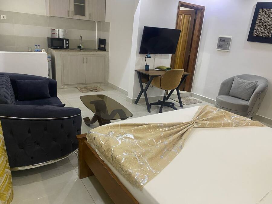 A compact hotel room with a bed, a sofa, a small desk with a chair, a TV, and a kitchenette.