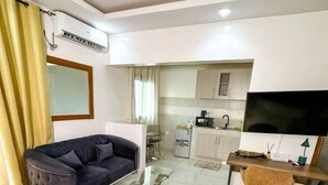 A hotel room with a bed, a sofa, a desk with a chair, a television, and a kitchenette in the background. - HOTL FABY 1 (Dakar)