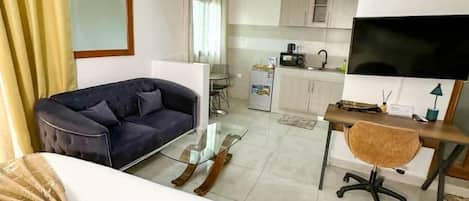 Habitación de hotel con cama, sofá, escritorio con silla, televisor y una kitchenette al fondo.