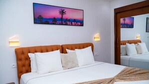 A hotel room with a bed, two wall-mounted lamps, a painting of a sunset, and a mirror. - HOTL FABY 1 (Dakar)