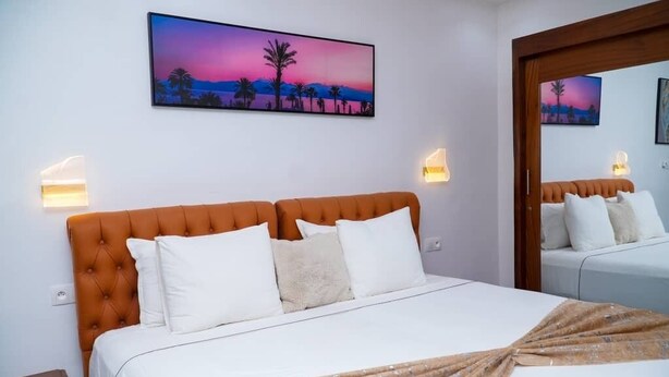 A hotel room with a bed, two wall-mounted lamps, a painting of a sunset, and a mirror. - HOTL FABY 1 (Dakar)