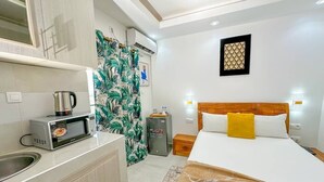 A hotel room with a bed, a small kitchenette, a shower curtain with a tropical pattern, a wall-mounted light fixture, and a framed artwork on the wall. - HOTL FABY 1 (Dakar)