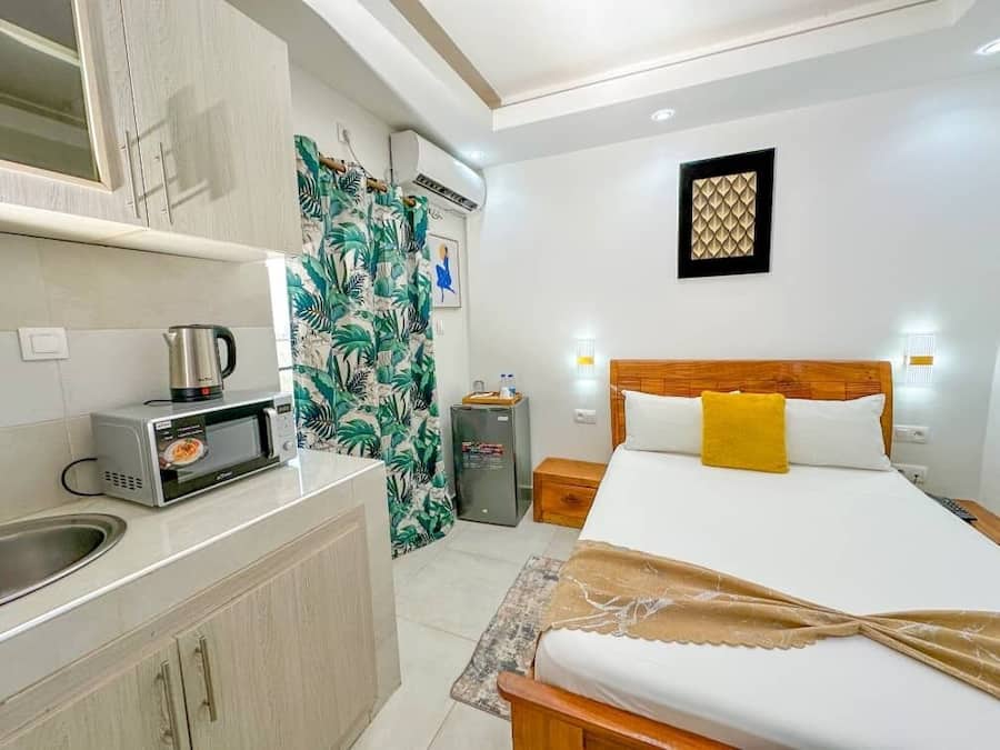 A hotel room with a bed, a small kitchenette, a shower curtain with a tropical pattern, a wall-mounted light fixture, and a framed artwork on the wall.
