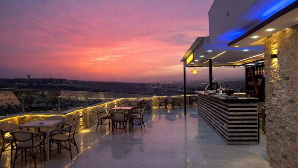 Une terrasse extérieure avec des tables et des chaises, un espace bar et une vue sur les toits de la ville au coucher du soleil.
