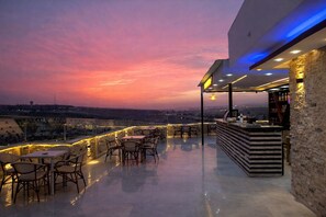 An outdoor terrace with tables and chairs, a bar area, and a view of the city skyline at sunset.