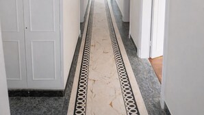 Un couloir aux murs blancs, doté d’un sol en marbre avec une bordure noire et blanche, et éclairé par un lustre suspendu.
