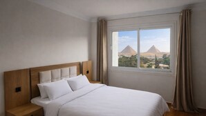 Une chambre à coucher avec une tête de lit en bois, du linge de lit blanc, une table de chevet et une fenêtre donnant sur les pyramides.