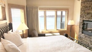 Habitación de hotel con una cama grande, chimenea de piedra, vista a un lago y un balcón con barandilla.