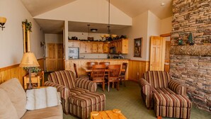 A cozy living room with striped armchairs, a sofa, and a wooden coffee table. A kitchen with wooden cabinets and a stone fireplace is visible in the background. - Lakeview Burlington Bay Two bedroom Suite (Two Harbors)