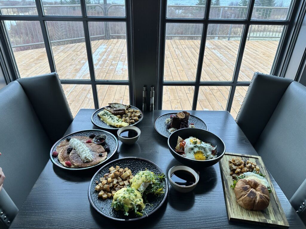 Uma mesa de jantar com diversos pratos, um deck de madeira visível através de grandes janelas e cadeiras estofadas pretas.