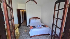 Ein schmales Hotelzimmer mit einem Bett, einem Deckenventilator und Blick auf einen Flur.
