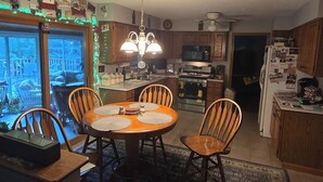 Una cocina con una mesa de comedor, sillas, una lámpara de araña, armarios de madera, un refrigerador y vistas a través de una puerta de cristal.