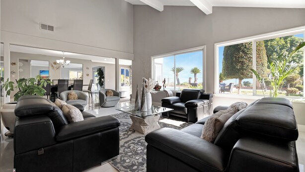 A spacious living room with black leather sofas, a glass coffee table, and large windows offering a view of palm trees and a clear sky.