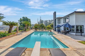 A rectangular swimming pool with a clear blue water surface, surrounded by a paved deck with lounge chairs and umbrellas.