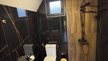 Un bany modern amb una paret revestida de rajoles negres i blanques, un moble de tocador de fusta, un vàter blanc, un petit tamboret de fusta i una zona de dutxa.
