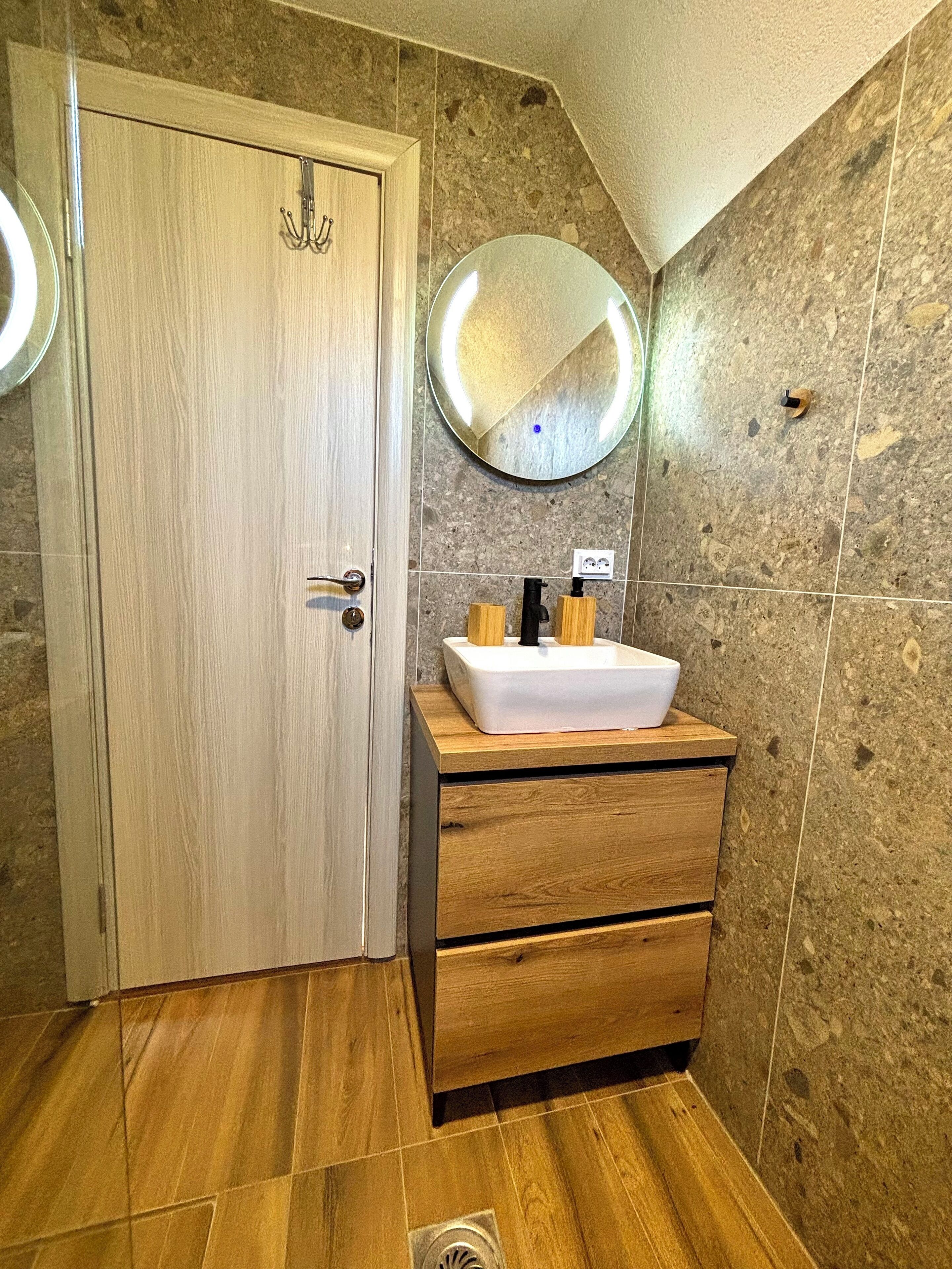 A bathroom with a wooden door, a round mirror, a white sink, and wooden drawers.