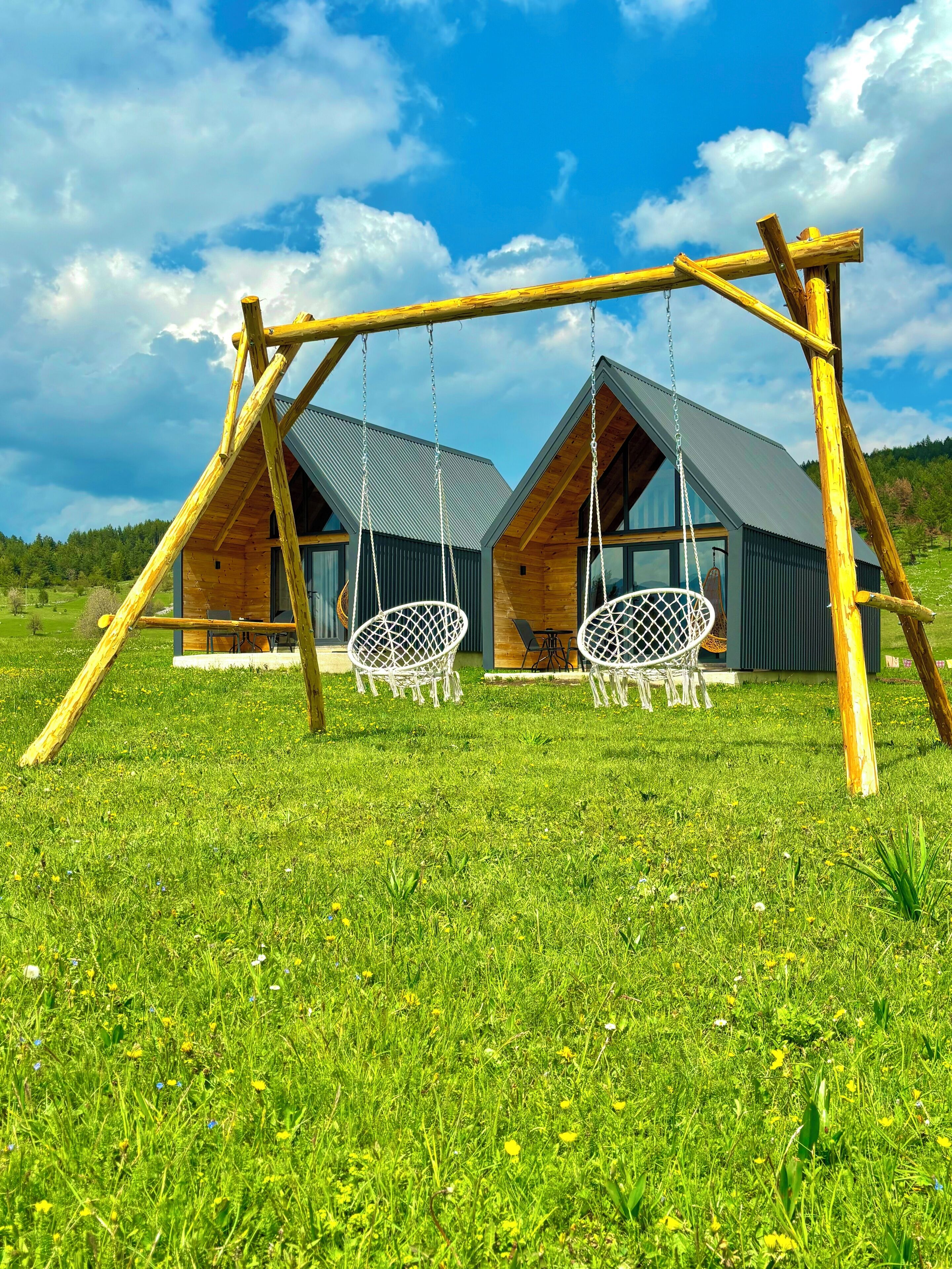 A wooden swing set with two swings in a grassy field, with two modern cabins in the background.