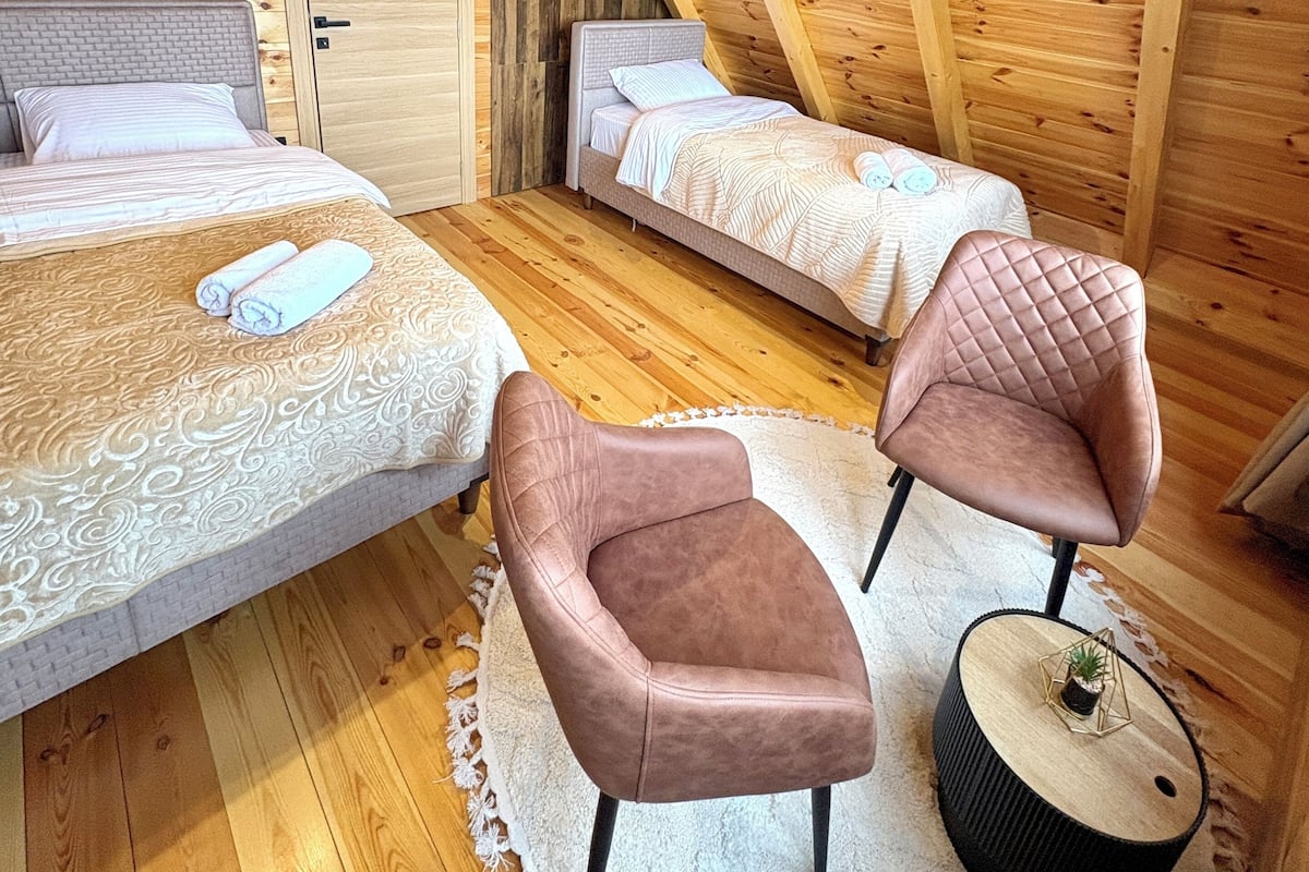 A cozy attic bedroom with wooden beams, two beds, a round coffee table, and a cushioned armchair.
