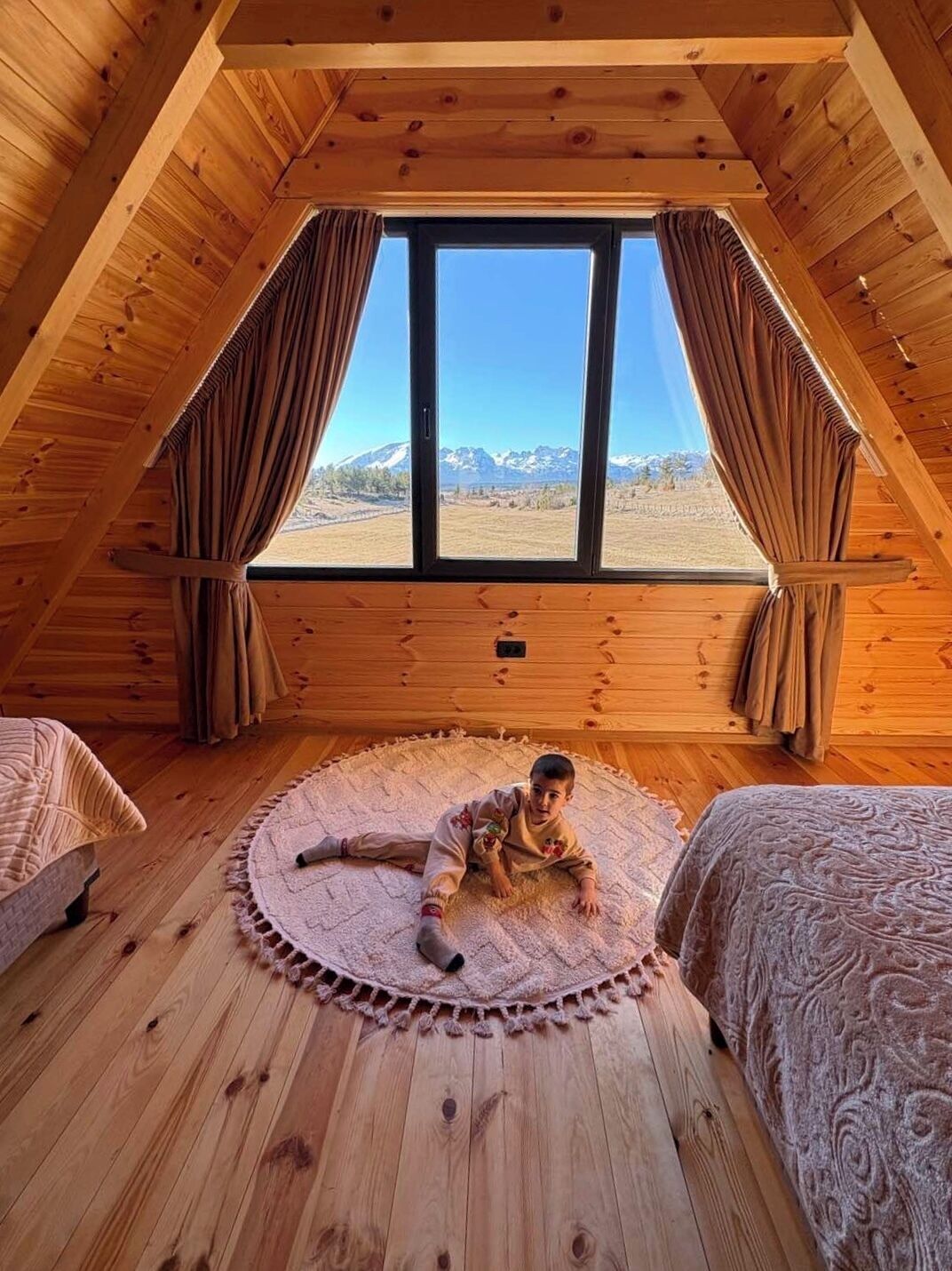 A cozy attic bedroom with a large window, wooden walls, and a child playing on a rug.