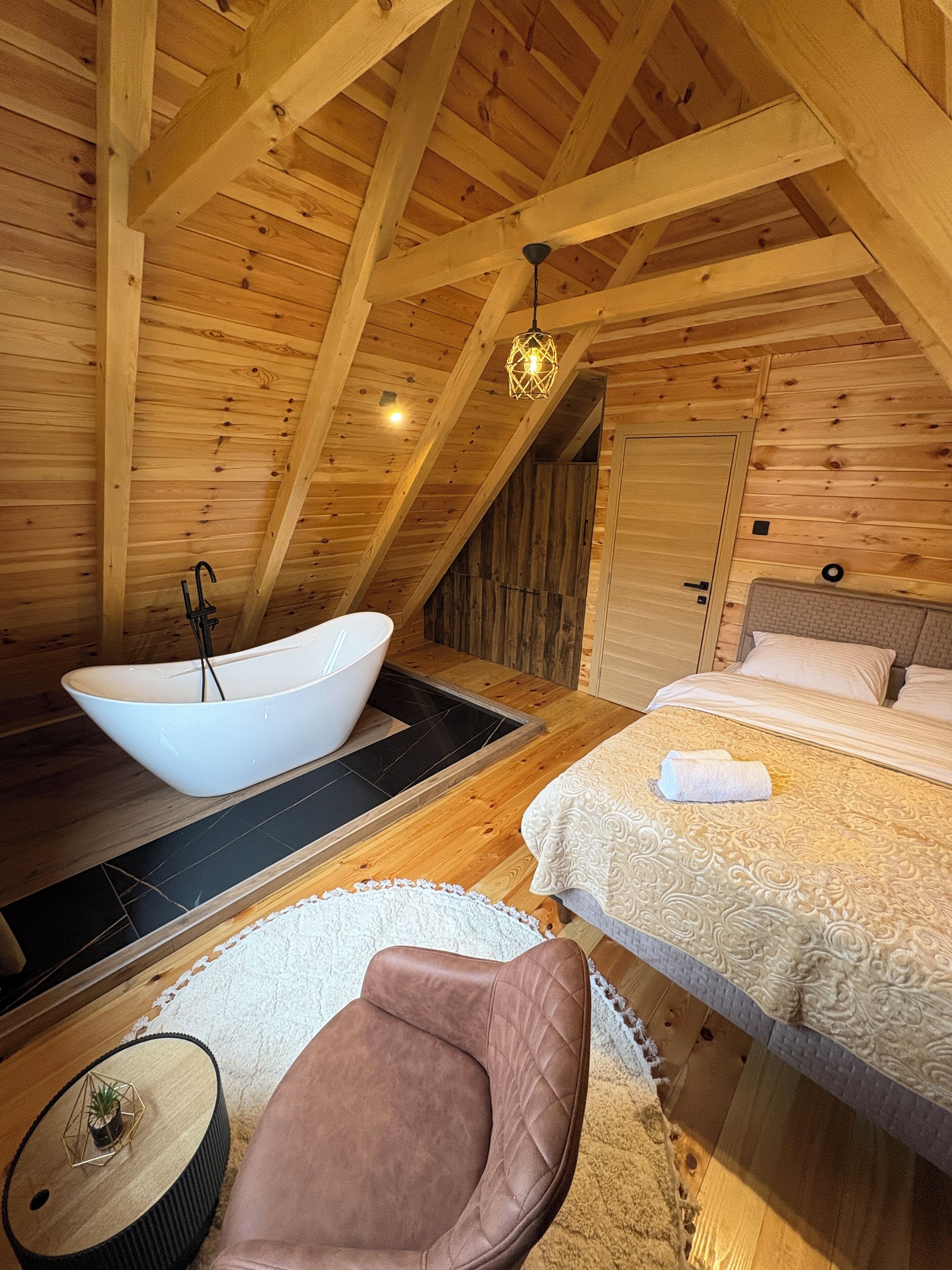 A wooden cabin interior with a large bathtub, a bed with a white bedspread, and a brown leather chair.