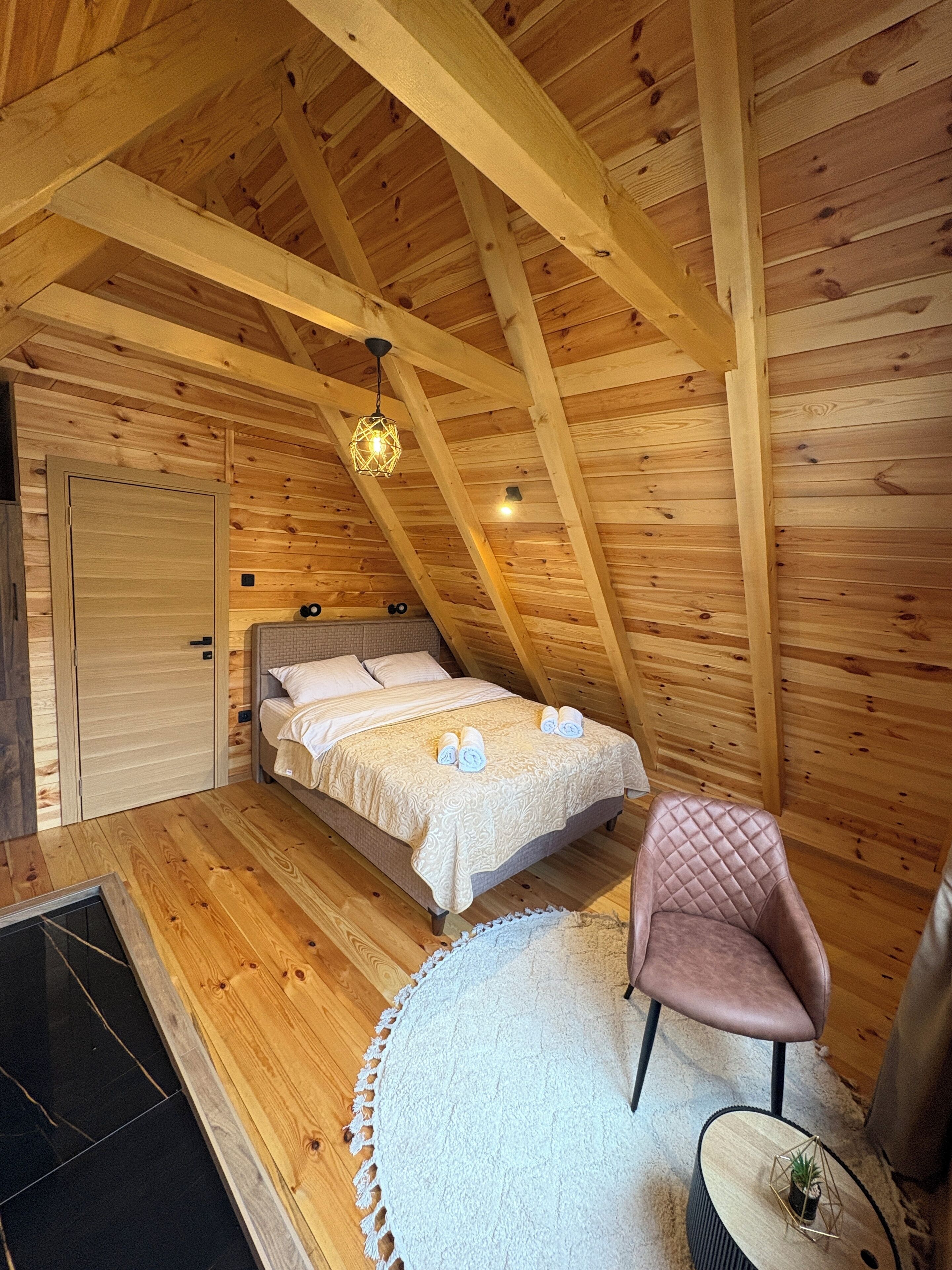 A wooden cabin bedroom with a bed, a chair, a small table with a plant, and a wooden ceiling with beams.