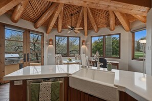 Une cuisine spacieuse dotée d’un grand îlot central, de poutres apparentes en bois au plafond et de grandes fenêtres offrant une vue sur l’extérieur.