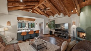 Amplia sala de estar con techo alto, vigas de madera, chimenea, televisor y zona de cocina con taburetes de bar.