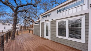 Une terrasse en bois avec une rambarde en métal, un grand arbre et un bâtiment à l’extérieur gris et aux ornements blancs.