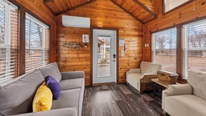 Interior acogedor de una cabaña con paredes de madera, un sofá gris, un sillón color beige y una pequeña mesa con un baúl antiguo.