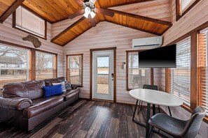 Un intérieur de chalet chaleureux avec un plafond en bois, un canapé en cuir agrémenté d’un coussin bleu, une table à manger avec une chaise, et un téléviseur à écran plat fixé au mur.