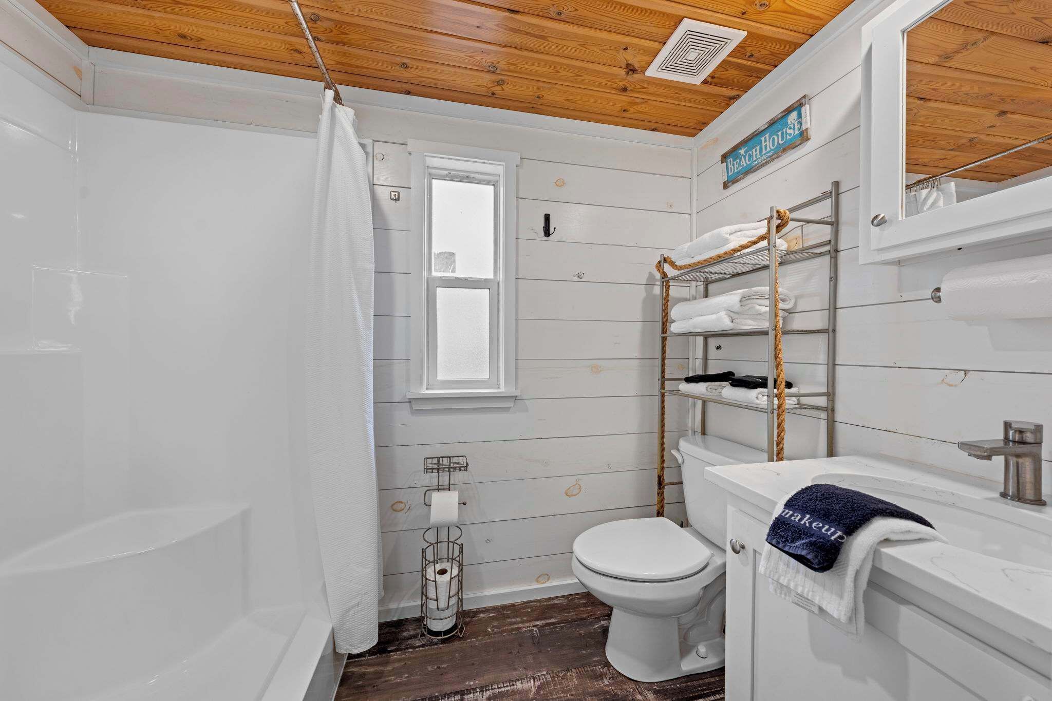 Un baño con techo de madera, una bañera blanca, cortina de ducha, inodoro y un perchero con toallas.