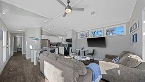 A modern living room with a grey sofa, a coffee table, and a TV. There is a kitchen in the background with white cabinets and a dining area with a small table and chairs. - The Resort at Fredericksburg Unit 29 (Fredericksburg)