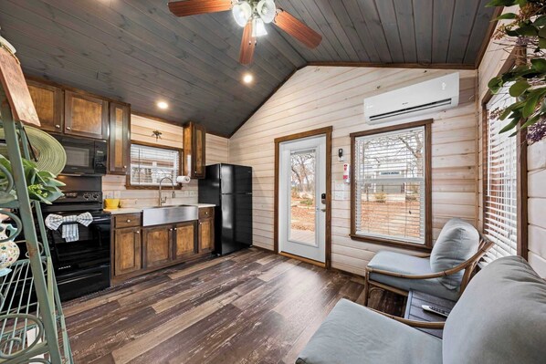 A compact kitchen with wooden cabinets, a microwave, stove, and refrigerator. There is a window with blinds, a ceiling fan, and a small seating area with a chair and cushion. - The Resort at Fredericksburg Unit 3 (Fredericksburg)