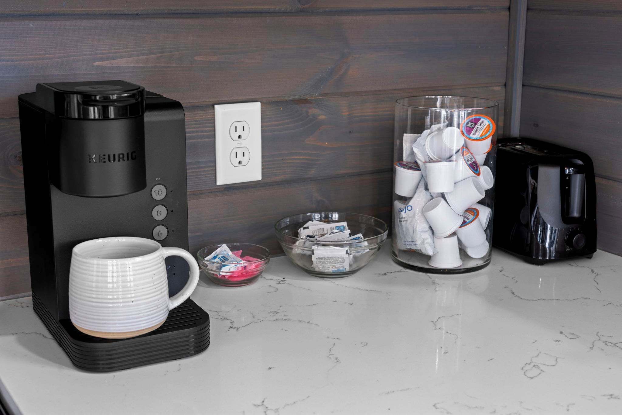 Una cafetera, un recipiente de vidrio con cápsulas de café y una taza sobre una encimera.