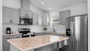 A modern kitchen with grey cabinets, a stainless steel refrigerator, a built-in microwave, a coffee station, and a large island with bar stools. - The Resort at Fredericksburg Unit 17 (Fredericksburg)