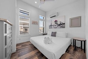 Une chambre à coucher avec un grand lit, du linge de lit blanc, un plancher en bois, une petite table de chevet et des œuvres d’art accrochées aux murs.