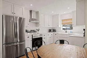 A modern kitchen with white cabinetry, stainless steel appliances, and a wooden dining table. - Das Morning Haus - Cozy and Spacious (Fredericksburg)